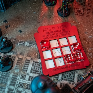 May include: Red wooden game board with the text "ACTS OF FAITH" and "GAIN ONE MIRACLE DICE AT THE START OF EACH TURN & WHEN YOUR UNIT IS DESTROYED". The board has nine squares with red dice in some of the squares. The board is on a grey and white surface.