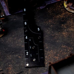 May include: Black measuring tool with white numbers and markings, used for tabletop gaming. The tool has a curved edge and a series of holes. The numbers range from 1 to 8. The tool is placed on a dark, textured surface.