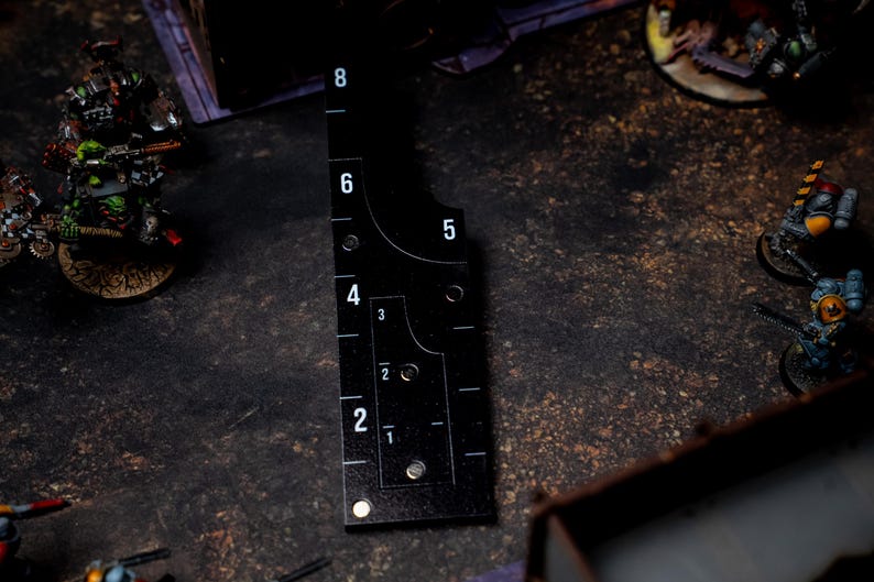 May include: Black measuring tool with white numbers and markings, used for tabletop gaming. The tool has a curved edge and a series of holes. The numbers range from 1 to 8. The tool is placed on a dark, textured surface.