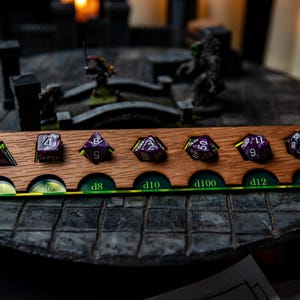 May include: A wooden dice tray with seven compartments for different sizes of dice. The tray has a green and brown color scheme and is labeled with the dice sizes: d4, d6, d8, d10, d100, d12, and d20.  The tray is filled with purple and black dice.