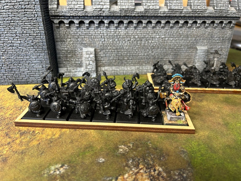 K&ouml;nnte beinhalten: Eine Gruppe von schwarz bemalten Miniaturfiguren von Kriegern mit &Auml;xten und Schilden, die auf einer Holzbasis stehen. Eine gr&ouml;&szlig;ere, detailliertere Miniaturfigur eines Kriegers mit einem Hammer und einem Schild, in Gold, Rot und Blau bemalt, steht vor der Gruppe.