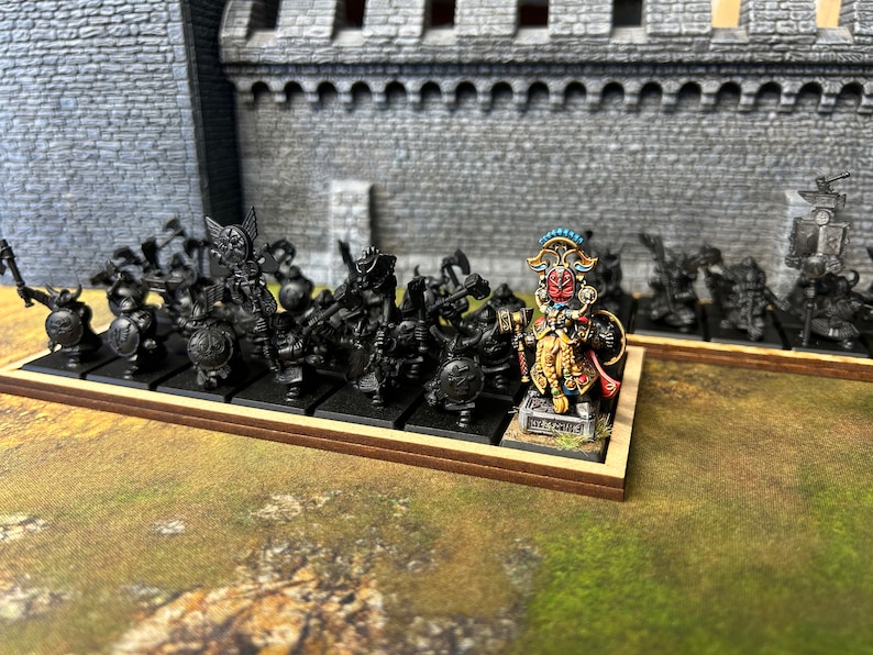 K&ouml;nnte beinhalten: Eine Miniatur-Kriegerarmee aus schwarz bemalten Figuren mit einer einzigen, gr&ouml;&szlig;eren, leuchtend bunten Figur in der Mitte. Die gr&ouml;&szlig;ere Figur tr&auml;gt ein goldenes und rotes Kost&uuml;m mit einem blauen und goldenen Kopfschmuck. Die Figuren stehen auf einem Holzsockel.