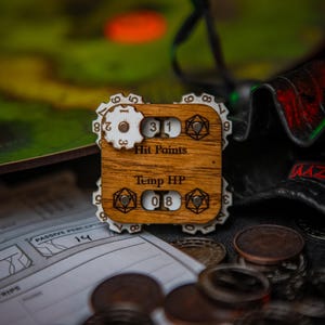 May include: A wooden character sheet tracker for tabletop role-playing games. The tracker has dials for hit points, temporary hit points, and other stats. The tracker is made of wood and has a natural finish. The tracker is sitting on a table with other game pieces.