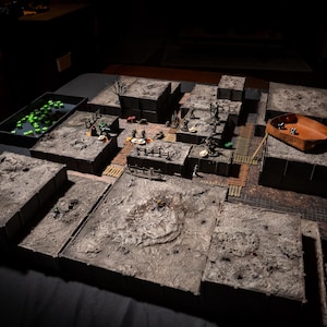 May include: A detailed tabletop role-playing game setup with modular terrain pieces. The scene includes miniature figures, dice, and a dice tray. The terrain is gray and brown, creating a realistic battleground for fantasy adventures.