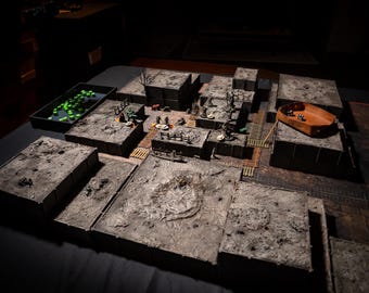 May include: A detailed tabletop role-playing game setup with modular terrain pieces. The scene includes miniature figures, dice, and a dice tray. The terrain is gray and brown, creating a realistic battleground for fantasy adventures.