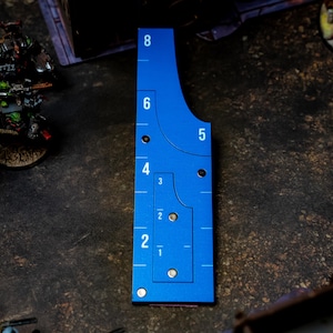 May include: A blue measuring tool with white markings for tabletop gaming. The tool has curved edges and numbered increments from 1 to 8 inches (approximately 20 cm). It is placed on a textured surface with miniature figures in the background.