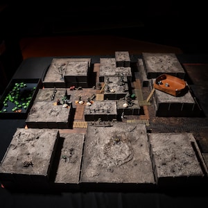 K&ouml;nnte beinhalten: Ein detailliertes Tabletop-Rollenspiel-Setup mit modularen Gel&auml;ndeteilen. Die Szene umfasst Miniaturfiguren, W&uuml;rfel und eine W&uuml;rfelschale aus Holz. Das Gel&auml;nde ist grau und braun und schafft ein realistisches Schlachtfeld.