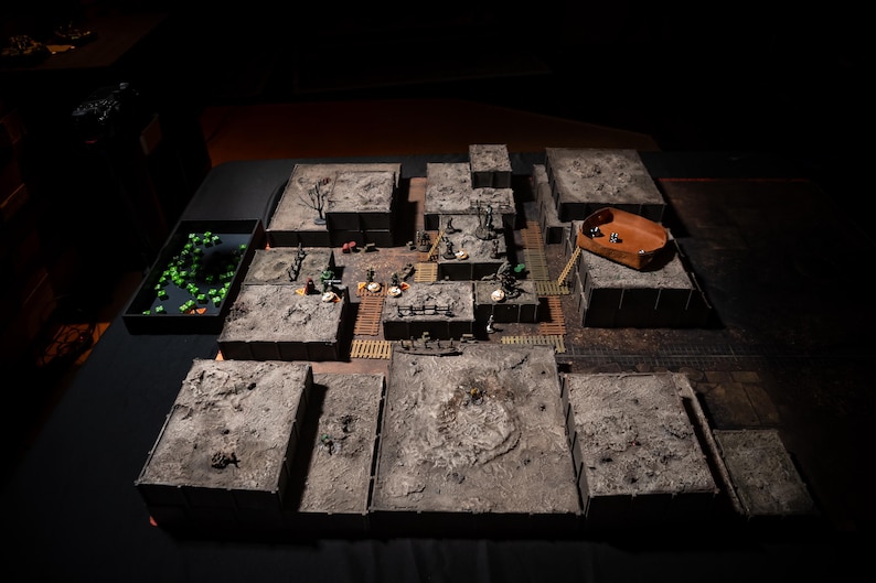 K&ouml;nnte beinhalten: Ein detailliertes Tabletop-Rollenspiel-Setup mit modularen Gel&auml;ndeteilen. Die Szene umfasst Miniaturfiguren, W&uuml;rfel und eine W&uuml;rfelschale aus Holz. Das Gel&auml;nde ist grau und braun und schafft ein realistisches Schlachtfeld.