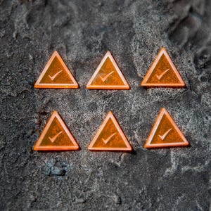 Może przedstawiać: Sześć pomarańczowych trójkątnych żetonów z białym symbolem znaku wyboru jest ułożonych na teksturowanej, ciemnoszarej powierzchni. Żetony mają półprzezroczysty wygląd, a znaki wyboru są dobrze widoczne.