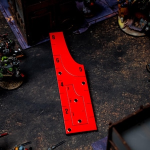 May include: A bright red gaming range ruler with black markings and numbers. The ruler has a curved design with multiple holes and is used for measuring distances in tabletop games. Miniature figures are visible in the background.