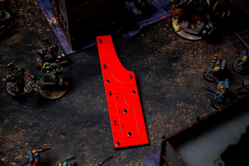 May include: A bright red gaming range ruler with black markings and numbers. The ruler has a curved design with multiple holes and is used for measuring distances in tabletop games. Miniature figures are visible in the background.
