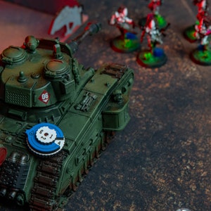 Op de afbeelding: Een groene militaire tank met een blauwe en witte ronde wijzerplaat aan de zijkant. De tank heeft het nummer "06" bovenop. De tank staat op een bruin oppervlak met verschillende miniatuurfiguren op de achtergrond.