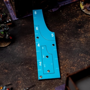 May include: A teal-colored game range ruler with black markings and numbers from 1 to 8. The ruler has a unique shape with curved edges and several circular holes. Miniature figures are visible in the background, suggesting a tabletop game.