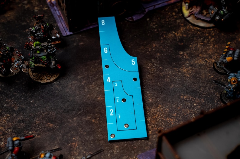 May include: A teal-colored game range ruler with black markings and numbers from 1 to 8. The ruler has a unique shape with curved edges and several circular holes. Miniature figures are visible in the background, suggesting a tabletop game.