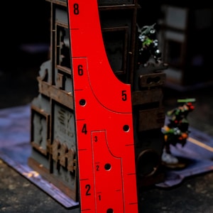 May include: A bright red game ruler with black markings and numbers, including 1 through 8, is displayed vertically. The ruler has a unique shape with rounded edges and several circular cutouts. The background features miniature game pieces and a building.