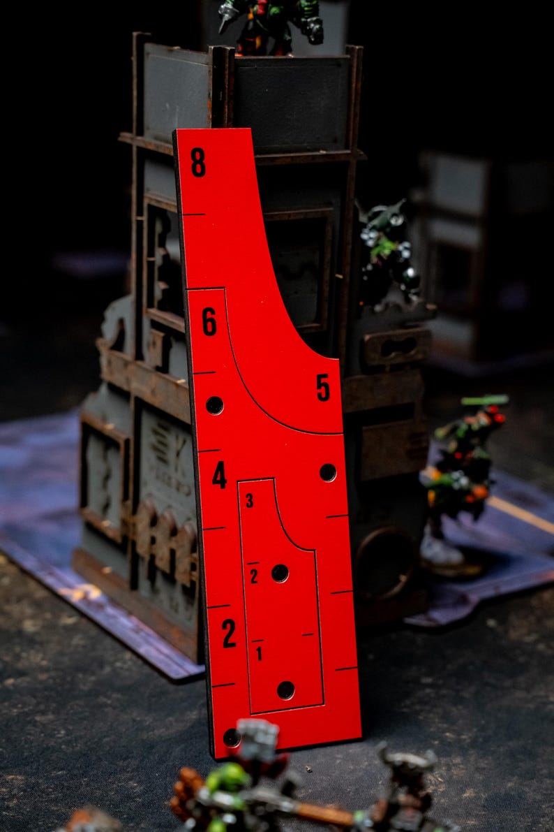 May include: A bright red game ruler with black markings and numbers, including 1 through 8, is displayed vertically. The ruler has a unique shape with rounded edges and several circular cutouts. The background features miniature game pieces and a building.