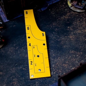May include: A yellow measuring tool with black markings and numbers, including 1 through 8, is shown. The tool has a unique shape with curved edges and is used for tabletop gaming. The tool is on a dark surface with miniature figures in the background.