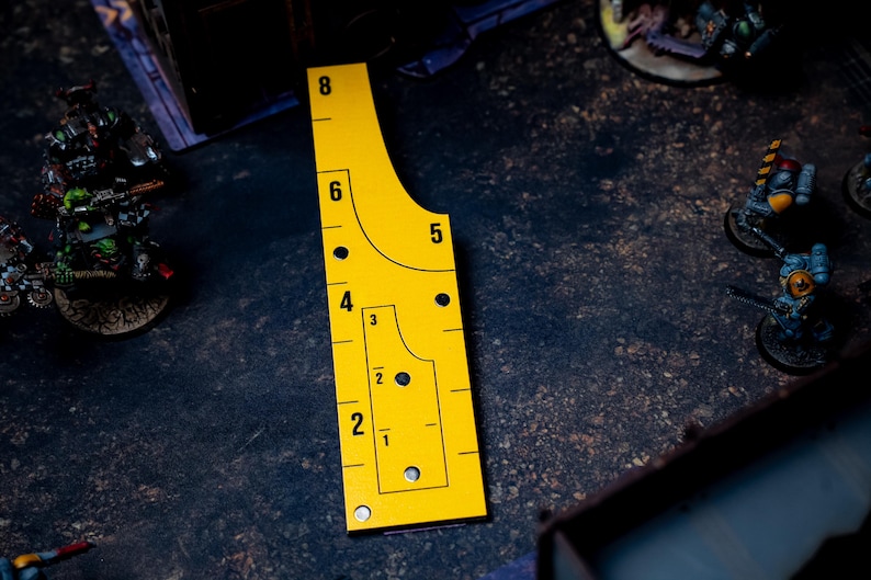 May include: A yellow measuring tool with black markings and numbers, including 1 through 8, is shown. The tool has a unique shape with curved edges and is used for tabletop gaming. The tool is on a dark surface with miniature figures in the background.