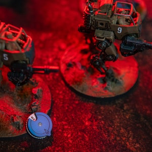 Op de afbeelding: Twee gedetailleerde, beschilderde, militaire miniatuurrobots op een rode achtergrond. De robots zijn gemaakt van metaal en plastic en hebben een futuristisch ontwerp. De robot links heeft het nummer 5 erop, en de robot rechts heeft het nummer 9 erop. Beide robots hebben kanonnen en staan op ronde bases.