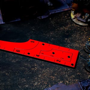 May include: A bright red game ruler with black markings and numbers, designed for tabletop gaming. The ruler has a curved edge and several holes, with miniature figures in the background.