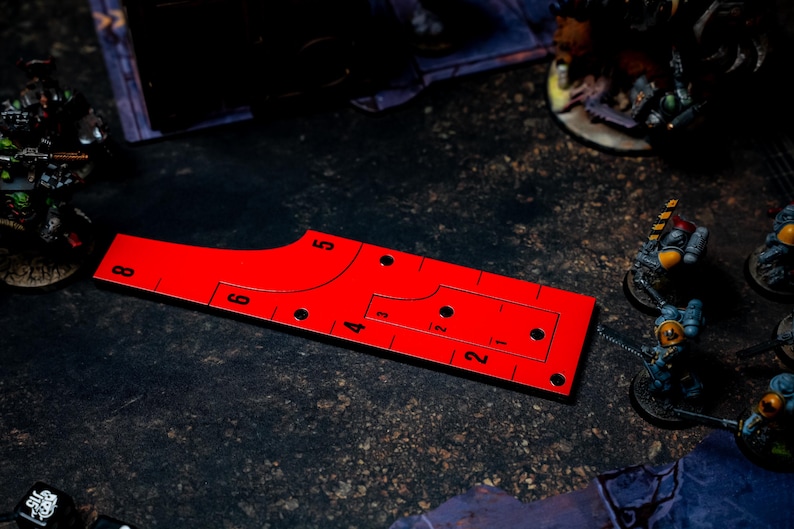 May include: A bright red game ruler with black markings and numbers, designed for tabletop gaming. The ruler has a curved edge and several holes, with miniature figures in the background.