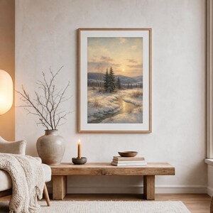 May include: A framed landscape print featuring a winter scene with a winding stream, snow-covered ground, and evergreen trees under a soft sunset sky. The artwork is displayed in a light wood frame hanging on a neutral wall above a wooden bench.