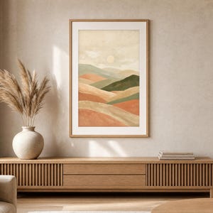 May include: A framed vertical wall art print featuring a landscape illustration of rolling hills in shades of terracotta, sage green, and beige under a pale sun. The artwork is enclosed in a light-toned wooden frame with a white mat border.