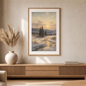May include: A framed vertical wall art print depicting a winter landscape with a snow-covered field, a winding stream, evergreen trees, and a soft sunset sky. The artwork is displayed in a light wood frame hanging on a neutral-toned wall.