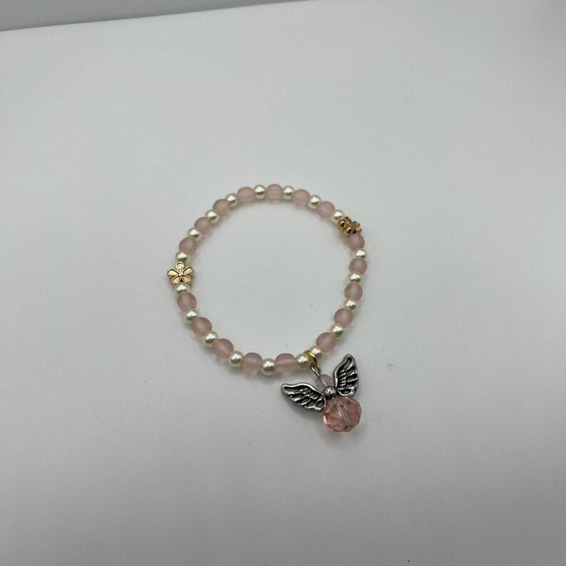May include: A beaded bracelet featuring pink and white beads, with a gold flower and a silver angel charm with a pink crystal. The bracelet is designed with elastic for easy wear.