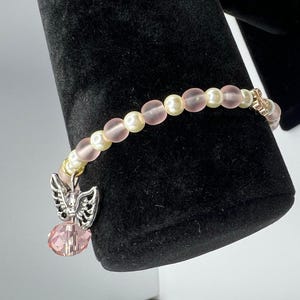 May include: A delicate bracelet featuring alternating pink and cream-colored beads. A silver-toned charm with an angel wing design and a pink crystal pendant hangs from the bracelet. The bracelet is displayed against a black background.
