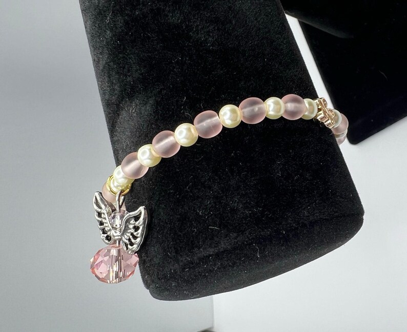 May include: A delicate bracelet featuring alternating pink and cream-colored beads. A silver-toned charm with an angel wing design and a pink crystal pendant hangs from the bracelet. The bracelet is displayed against a black background.