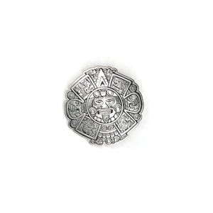 May include: A circular silver-toned metal medallion featuring a detailed relief of the Aztec sun stone calendar. The design includes a central face with a protruding tongue surrounded by concentric rings of intricate glyphs and symbols.