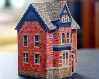May include: A red brick paper model of a two-story house with a blue roof and trim. The house has windows and a door.