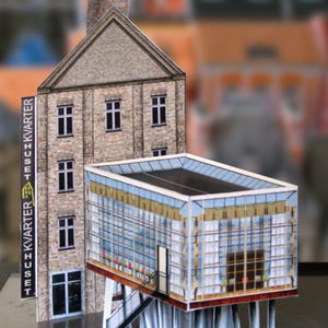May include: A paper model depicting two buildings. One is a brick structure with a chimney and a sign reading "KVARTER". The other is a modern glass building raised on white supports. The buildings are set against a blurred backdrop.
