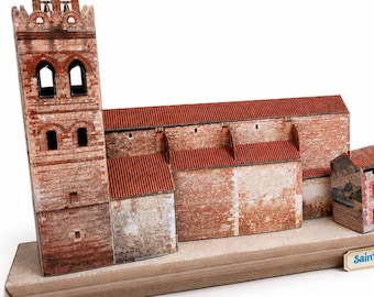 May include: A paper model of a church with a tall bell tower and red tiled roofs, featuring printed brick and stone textures. The structure is mounted on a rectangular base and includes a small sign with the text Saint-Cyprien visible.