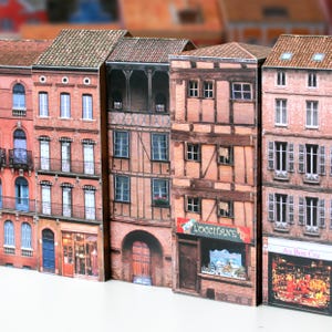 Puede incluir: Una fila de cinco edificios de ladrillo en miniatura con diferentes estilos arquitectónicos. Los edificios tienen fachadas de ladrillo marrón, rojo y blanco con ventanas y puertas. Los edificios tienen letreros que dicen "L'Occitane" y "Au Bon Cru".