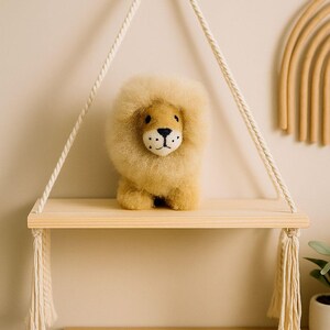 May include: A plush lion toy with a fluffy mane sits on a wooden shelf suspended by rope. The lion has a tan body, a light brown mane, and a detailed face. The shelf is part of a macrame wall hanging, adding a touch of bohemian decor.