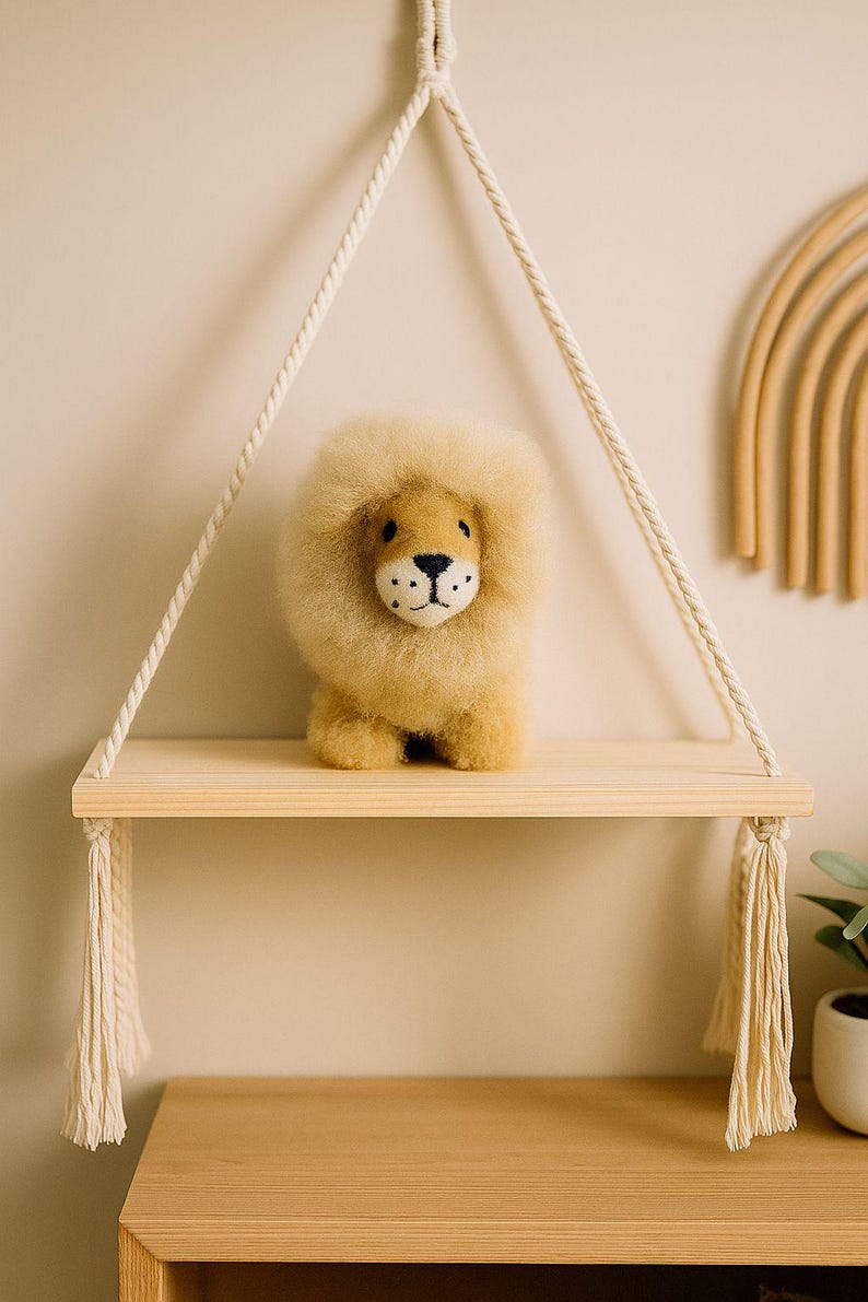 May include: A plush lion toy with a fluffy mane sits on a wooden shelf suspended by rope. The lion has a tan body, a light brown mane, and a detailed face. The shelf is part of a macrame wall hanging, adding a touch of bohemian decor.