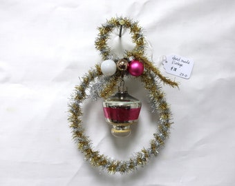May include: A handmade vintage Christmas ornament shaped like a snowman. The snowman is made of gold and silver tinsel and has a pink and white glass ball ornament hanging from it. The ornament has a tag that says "Hand made Vintage $1.00 23-D".