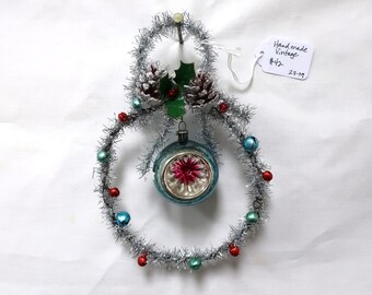 May include: A silver tinsel Christmas wreath with red and blue bells and a blue glass ornament with a pink star design. The wreath is decorated with two pine cones and a sprig of holly.