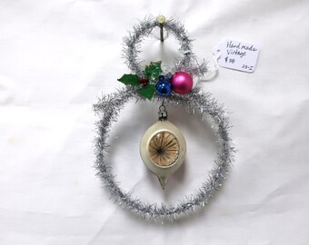 May include: Handmade vintage snowman ornament made of silver tinsel with a white glass bulb ornament hanging in the center. The snowman has a green holly sprig with red berries and blue and pink glass ball ornaments.