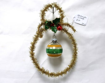 May include: A handmade vintage snowman ornament made of gold tinsel with a green and yellow striped glass ornament hanging from it. The snowman has a green holly sprig with red berries on top. The ornament has a handwritten tag that says "Handmade Vintage #30 23-N".