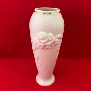 May include: A white porcelain vase with a gold rim and a raised design of a single rose. The vase is on a red background.