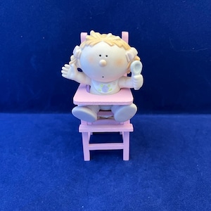 May include: A pink high chair with a small figurine of a baby girl sitting in it. The baby is holding a spoon and has blonde hair.