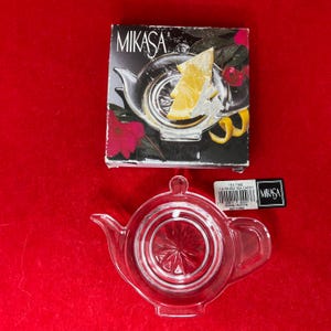 May include: A clear glass tea caddy in the shape of a teapot, with a matching box featuring a lemon slice and the Mikasa brand name. The caddy has a handle and spout, and the box is on a red surface.