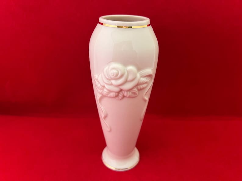 May include: A white porcelain vase with a gold rim and a raised design of a single rose. The vase is on a red background.