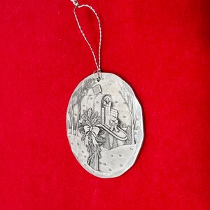 May include: Silver-colored Christmas ornament with a winter scene. The oval ornament features a mailbox with gifts, a bow, and a sprig of holly. The background shows trees and falling snow. A silver string is attached for hanging.