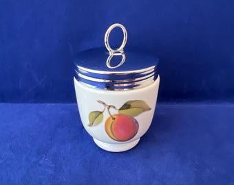 May include: A white ceramic egg cup with a silver lid and a painted image of a peach. The egg cup has a silver handle on the lid.