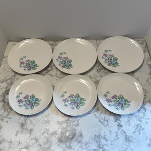 May include: Six white ceramic plates with a floral design. Each plate features a cluster of purple, blue, and green flowers in the corner. The plates are arranged on a light-colored surface.