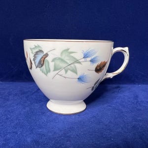 May include: White teacup with a floral design featuring blue, green, and brown leaves and flowers. The rim and handle are trimmed with gold. The teacup sits on a small, white pedestal base.